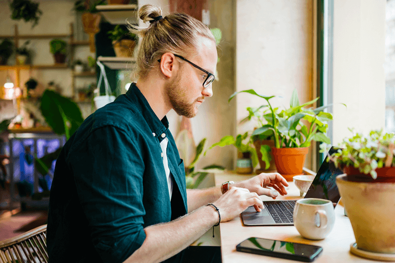 a man with glasses and blond hair works on a laptop with a mug and plants