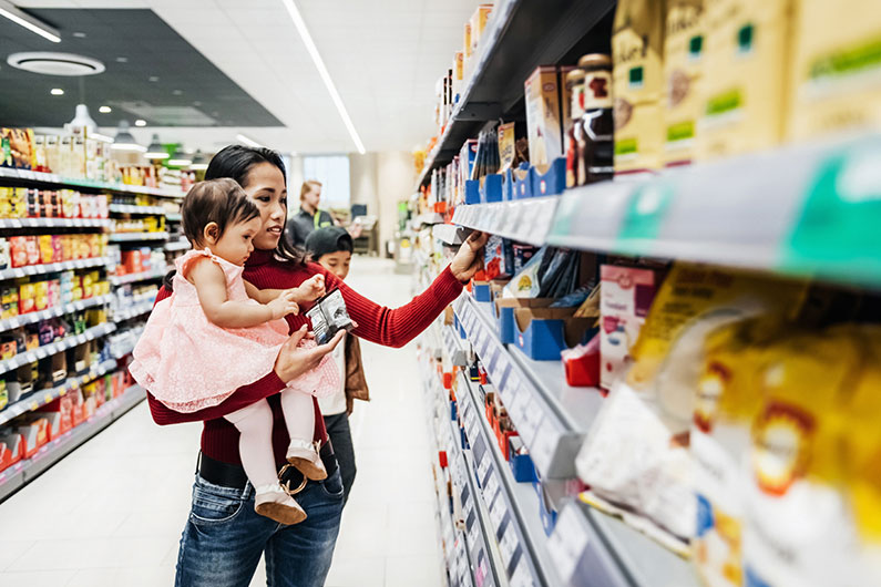 A young woman wearing a red sweater holds her toddler daughter while picking out snacks in a brightly lit grocery store.