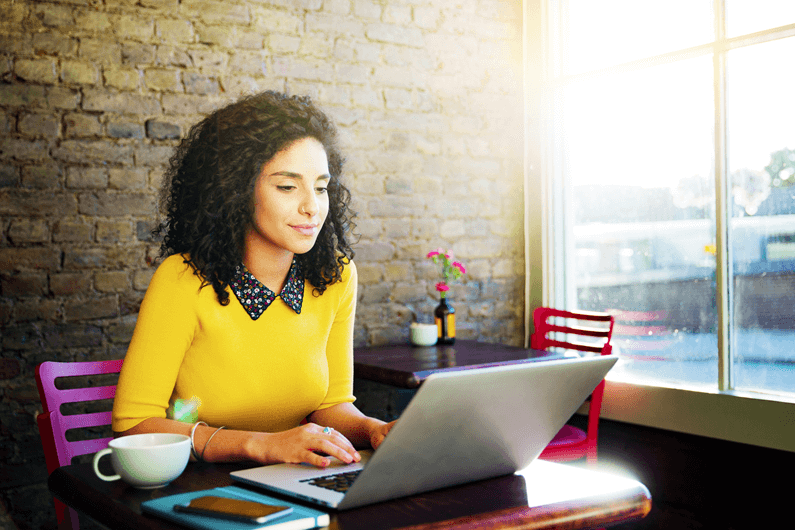 a woman in a yellow sweater smiles as she works on a laptop in a coffee shop